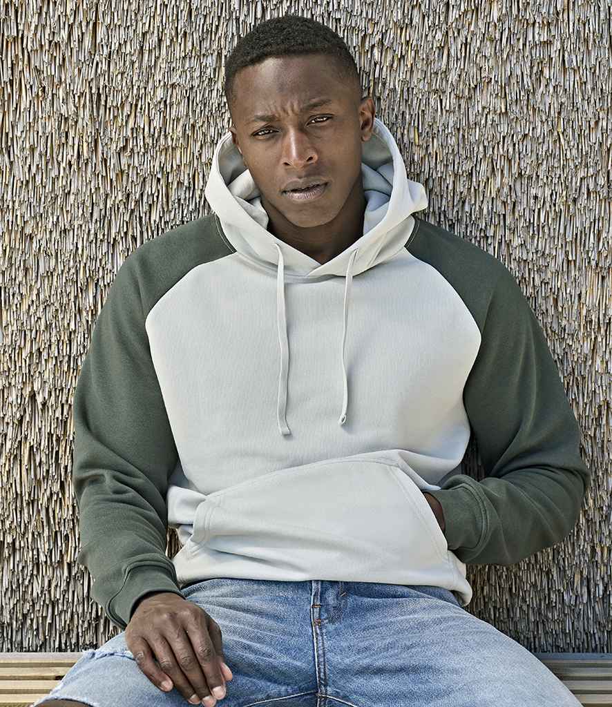 A man in a Tee Jays Unlabeled Two-Tone Hooded Sweatshirt with green sleeves and a light gray body sits by a textured wall. The tag-free, brushed back fleece offers comfort, complementing his laid-back style with ripped blue jeans.