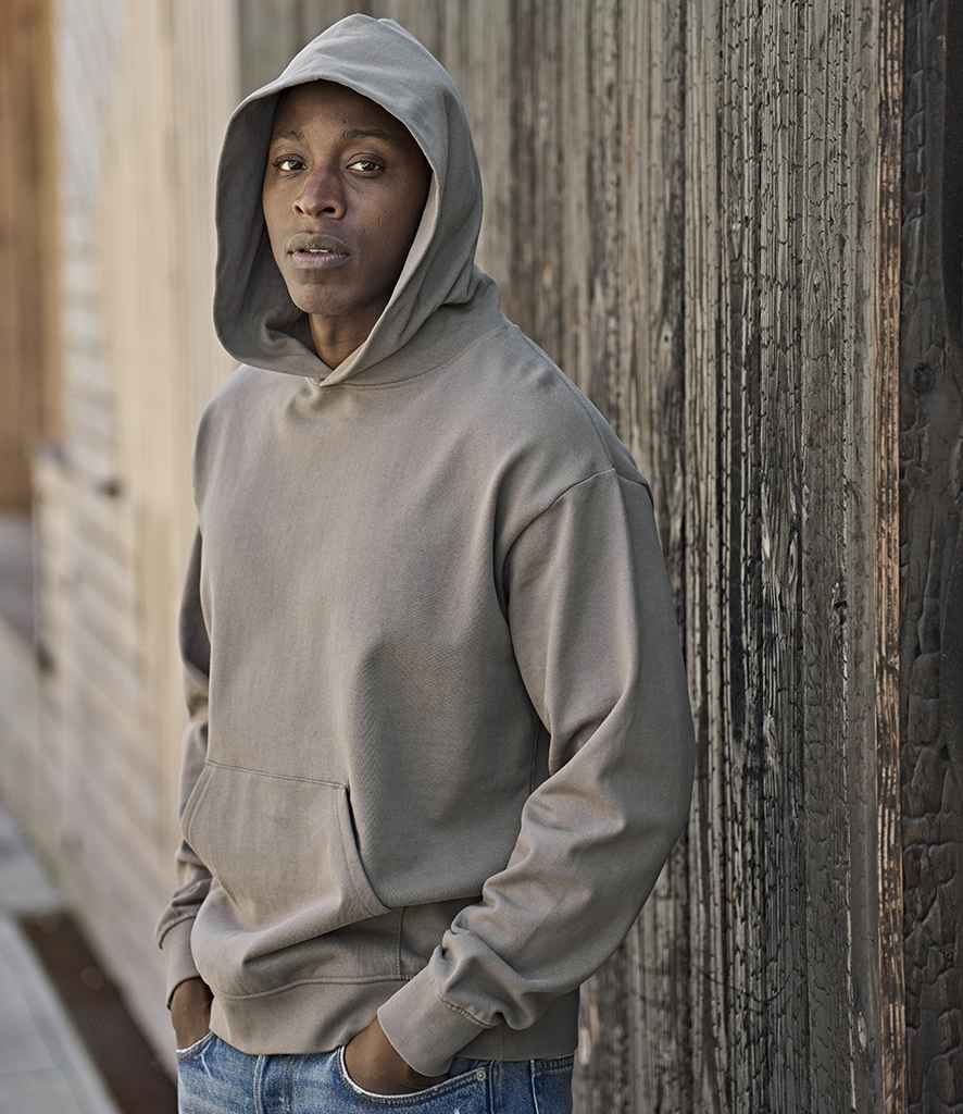 A person wears a Tee Jays Unlabeled Heavy Loose Fit Hooded Sweatshirt in gray with the hood up and hands in the front pocket, paired with blue jeans, standing against a wooden wall and looking at the camera neutrally.
