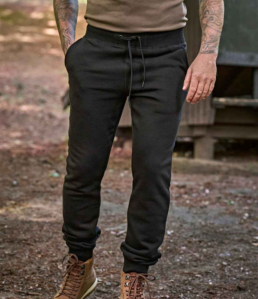 Outdoors, a person wears Tee Jays Unisex Sweat Pants with side pockets, a brown shirt, and hiking boots. Hands rest in the sweat pant pockets, tattoos show on their arms, and dirt and leaves cover the ground.