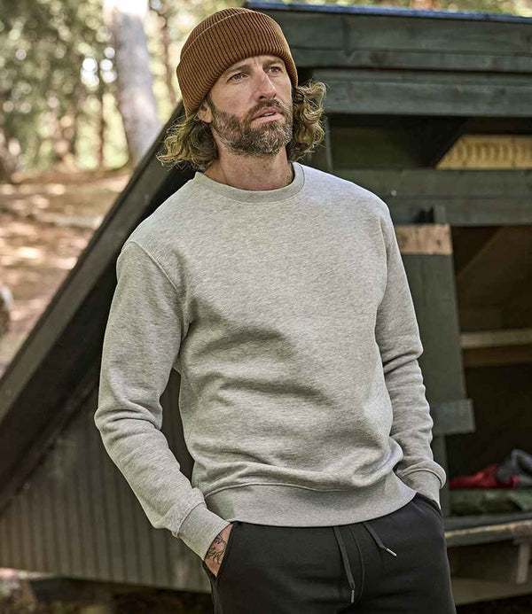 A bearded man wears a brown knit beanie and a light gray Tee Jays Heavy Sweatshirt by Tee Jays in cozy brushed back fleece, standing outdoors near a wooden structure with trees in the background.