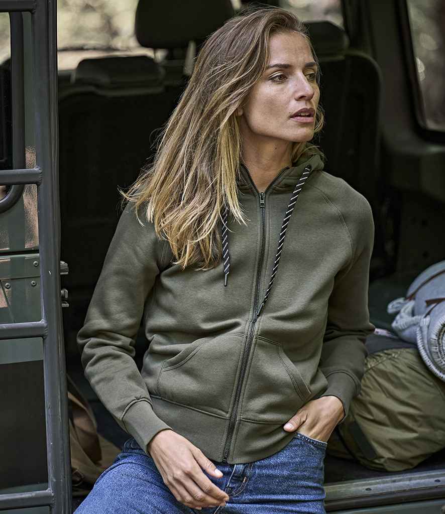 A woman with long blonde hair, wearing a Tee Jays Ladies Fashion Zip Hooded Sweatshirt in green and blue jeans, sits in the open doorway of a vehicle, looking to the side. A backpack and gear are visible inside.
