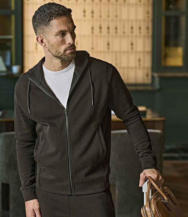 A man with short dark hair and a beard wears a Tee Jays Ribbed Interlock Full Zip Hooded Sweatshirt in black over a white t-shirt, holding the handle of a brown leather bag and looking to his left indoors.