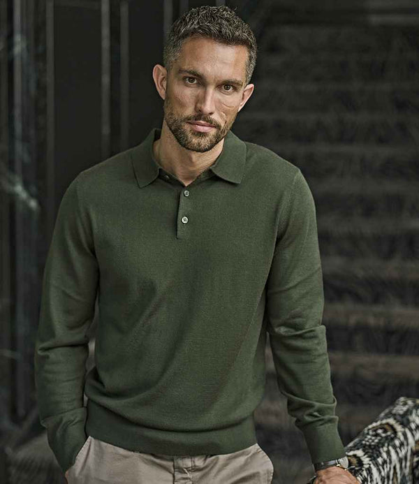A man with short hair and a beard stands indoors, wearing a dark green Tee Jays Knitted Polo Shirt with a button placket and light-colored pants. He looks at the camera with a neutral expression, one hand in his pocket.