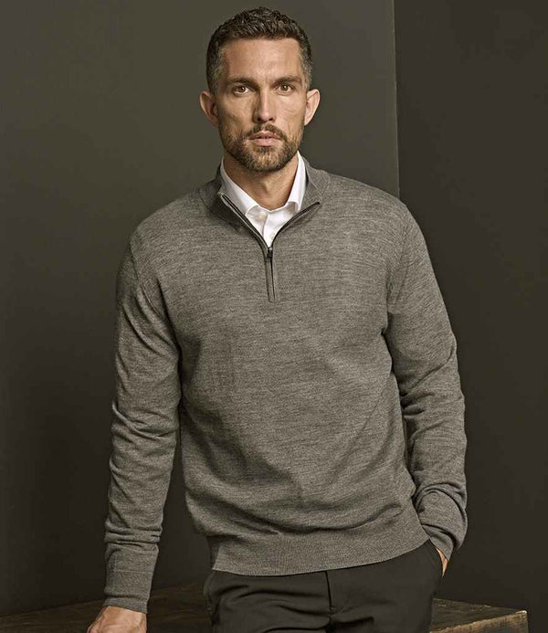 A man with short brown hair and a trimmed beard wears a Tee Jays Half Zip Sweater in gray, layered over a white collared shirt. He stands against a dark wall, leaning one hand on a wooden surface.