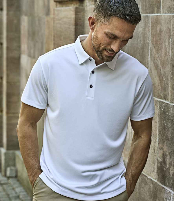 A man with short hair and a trimmed beard stands outdoors against a stone wall, looking down with his hands in his pockets. He wears the Tee Jays Luxury Sport Polo Shirt for all-day comfort, paired with beige pants.