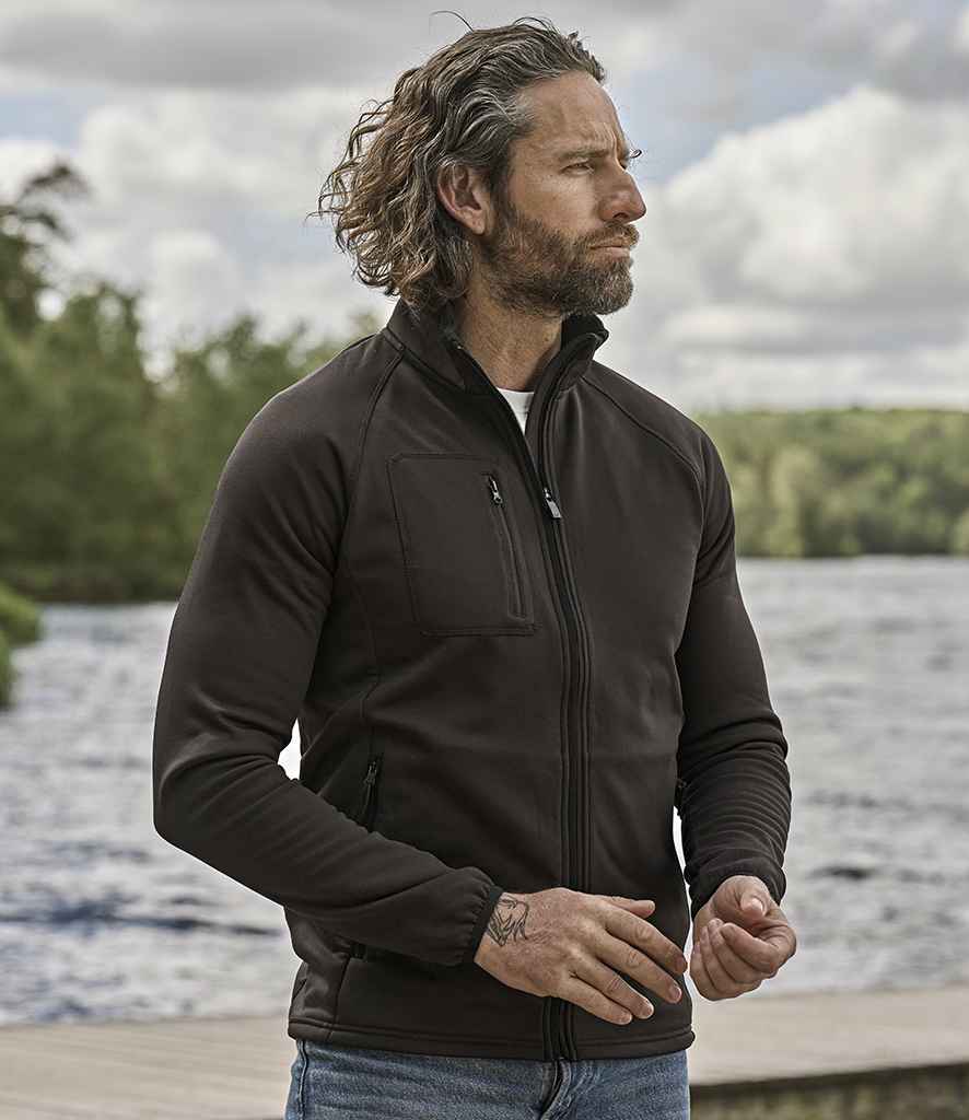 A man with wavy hair and a beard stands outdoors by water, wearing the Tee Jays Stretch Fleece Jacket from Tee Jays, featuring a dark zip-up design with a stand-up collar and jeans. Trees and a cloudy sky can be seen in the background.