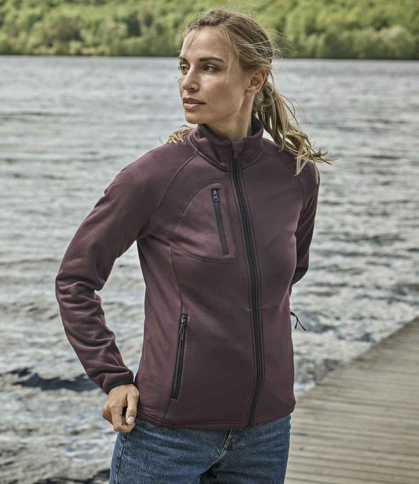 A woman in a Tee Jays Ladies Stretch Fleece Jacket, dark purple with a tailored fit, and blue jeans stands on a wooden dock by the water with green trees behind her. She looks slightly to her left.
