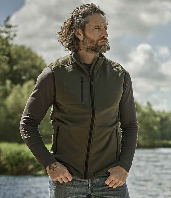 A man with wavy medium-length hair and a beard stands outdoors near water, wearing the Tee Jays Stretch Fleece Bodywarmer over a long-sleeve shirt and jeans. Trees and a cloudy sky are in the background.