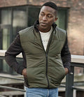 A man stands outdoors by a metal railing, wearing the Tee Jays Hybrid-Stretch Jacket over a white T-shirt and blue jeans, with a modern building featuring large windows in the background.