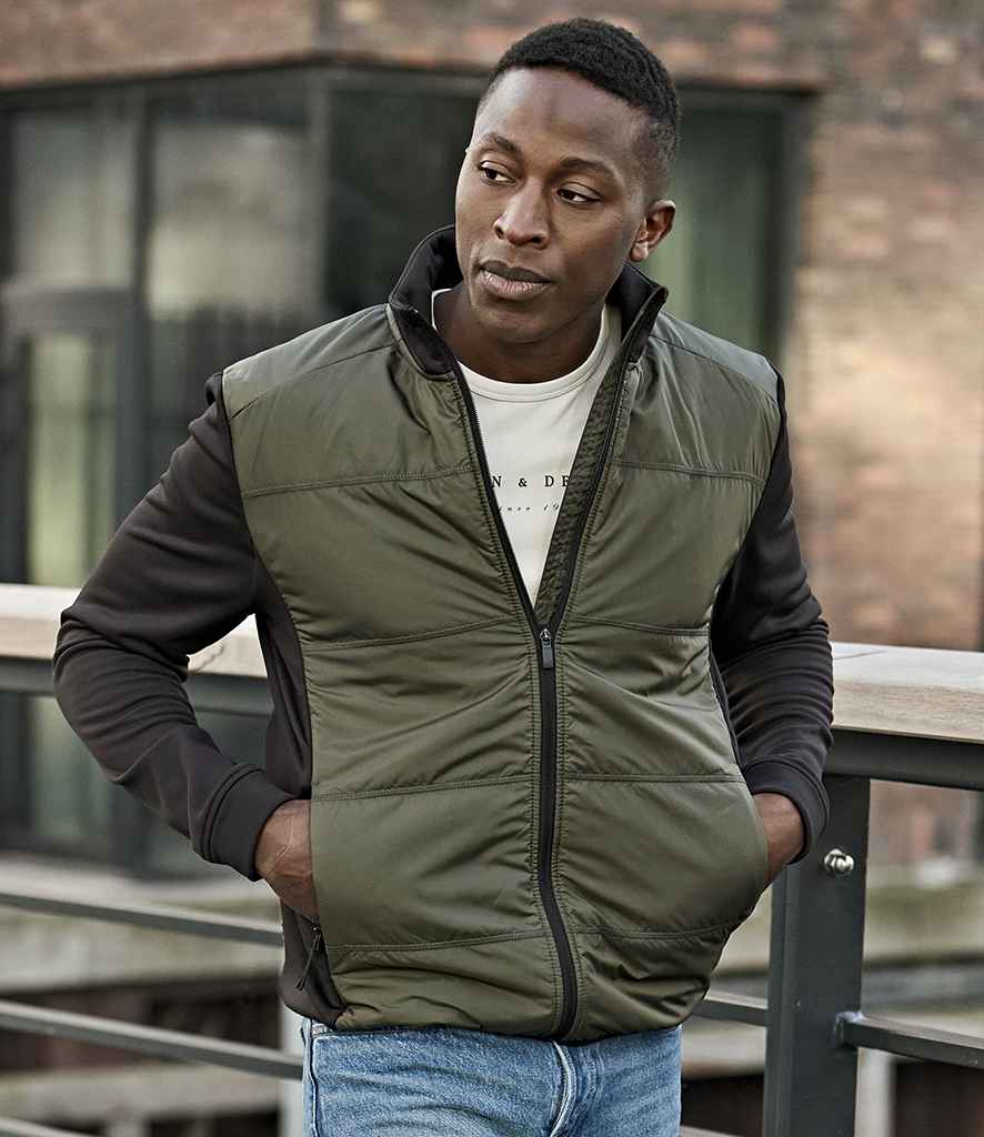 A man stands outdoors by a metal railing, wearing the Tee Jays Hybrid-Stretch Jacket over a white T-shirt and blue jeans, with a modern building featuring large windows in the background.