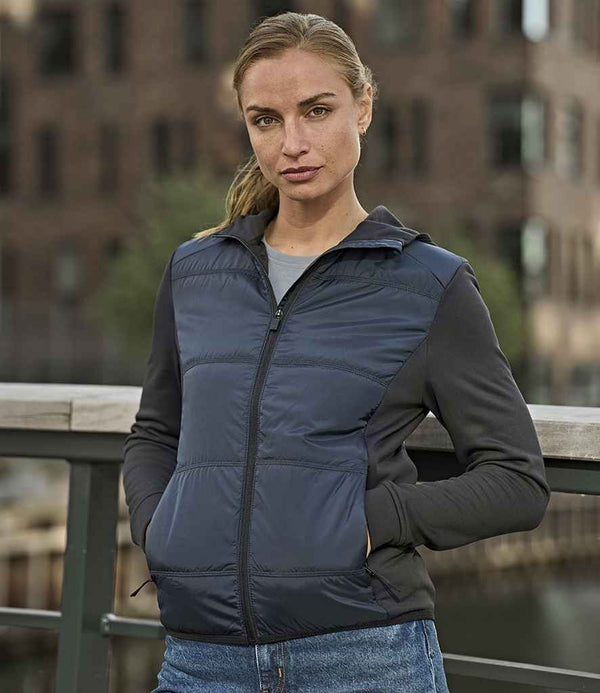 A woman stands outdoors in front of a brick building and railing, wearing the Tee Jays Ladies Hybrid-Stretch Hooded Jacket in navy and black with blue jeans. She faces the camera with a neutral expression.