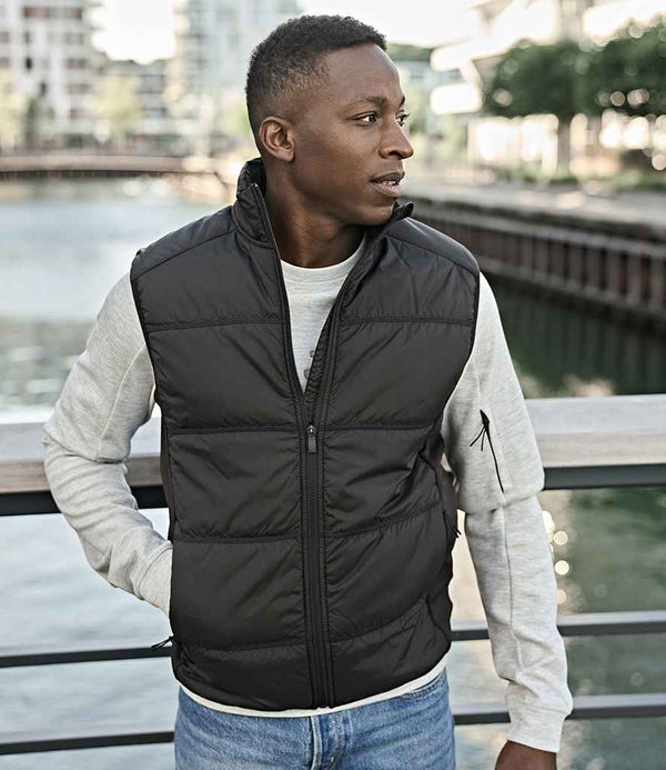 A man stands outdoors by a waterfront railing, wearing the Tee Jays Hybrid-Stretch Bodywarmer—a black sleeveless puffer vest by Tee Jays—over a light gray long-sleeve shirt and blue jeans. He looks left with one hand in his pocket.