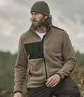 A man with wavy hair and a beard wears the olive green Tee Jays Mountain Fleece Jacket by Tee Jays, along with a dark knit beanie and dark pants. He is seated outdoors with a blurred natural landscape in the background.