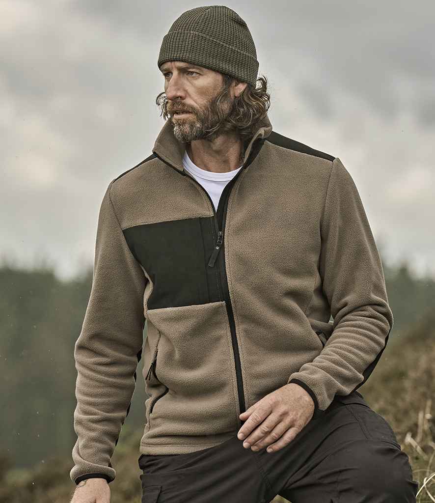 A man with wavy hair and a beard wears the olive green Tee Jays Mountain Fleece Jacket by Tee Jays, along with a dark knit beanie and dark pants. He is seated outdoors with a blurred natural landscape in the background.