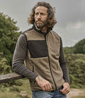 A man with wavy, shoulder-length hair and a beard stands outdoors by a wooden fence, wearing the Tee Jays Mountain Fleece Bodywarmer—a beige and black zip-up vest with chest pocket and reinforced ripstop polyester—over a dark shirt and blue jeans.