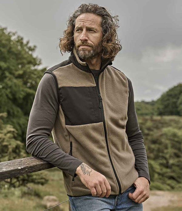 A man with wavy, shoulder-length hair and a beard stands outdoors by a wooden fence, wearing the Tee Jays Mountain Fleece Bodywarmer—a beige and black zip-up vest with chest pocket and reinforced ripstop polyester—over a dark shirt and blue jeans.