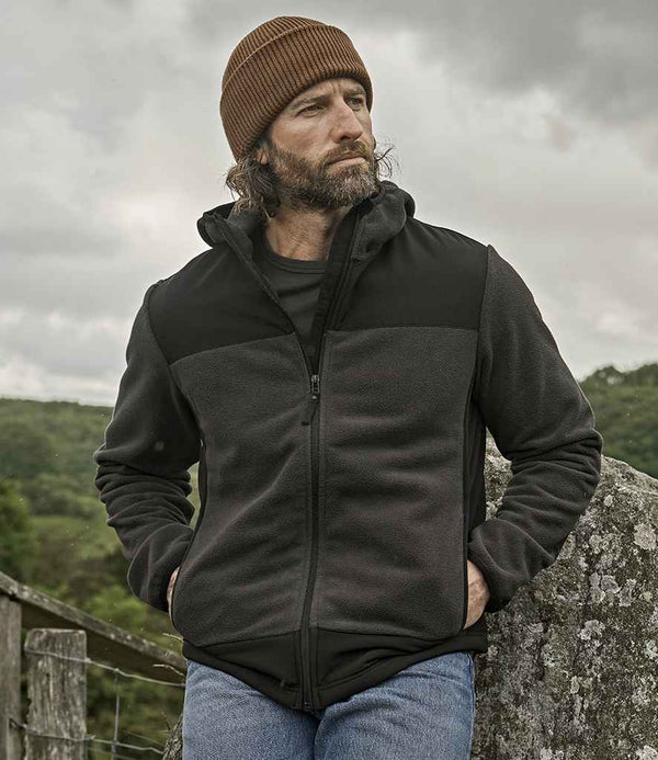 A man with a beard and long hair, wearing a brown knit beanie, blue jeans, and a Tee Jays Mountain Hooded Fleece Jacket by Tee Jays, stands outdoors against a stone wall with a cloudy sky and green landscape in the background.