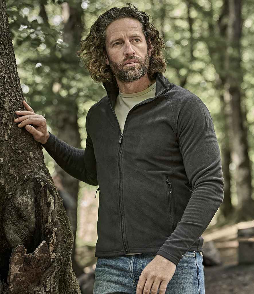 A man with long curly hair and a beard wears a Tee Jays Active Fleece Jacket and jeans, standing outdoors in a wooded area with one hand resting on a tree trunk.