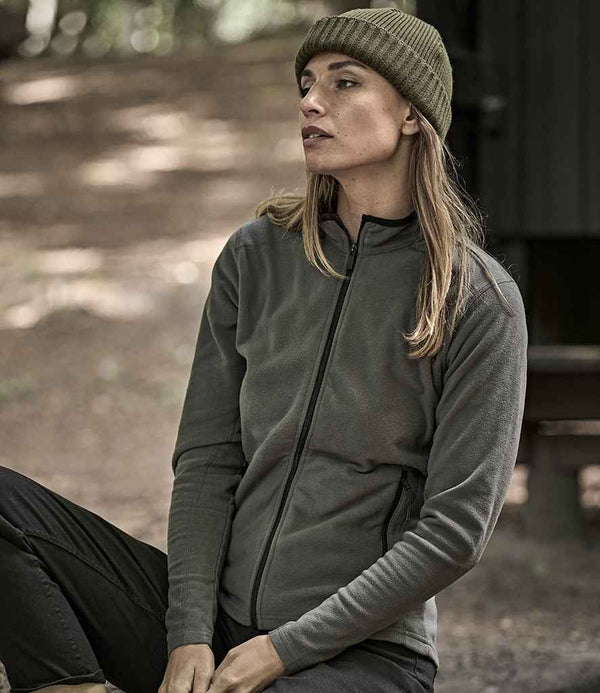 A person in a Tee Jays Ladies Active Fleece Jacket and green beanie sits on a wooden bench outdoors, looking to the side with trees and dappled sunlight in the background.