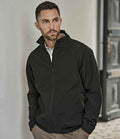 A man with short dark hair and a trimmed beard wears the Tee Jays Club Jacket by Tee Jays— a black, showerproof zip-up—over a white shirt. He stands indoors, leaning against a white wall and gazes to the side neutrally.