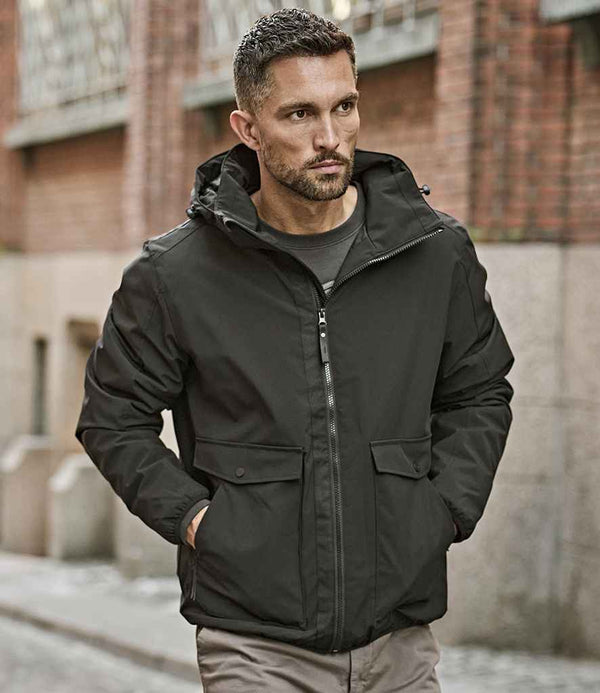 A man with short hair and a beard is wearing the Tee Jays Urban Adventure Shell Jacket by Tee Jays, a black windproof jacket with front pockets and a zipper, while standing outdoors on a city street near a brick building.