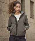 A person with curly hair wears the Tee Jays Ladies Urban Adventure Shell Jacket by Tee Jays in gray over a white shirt and dark pants, standing hands-in-pockets before a textured stone wall.