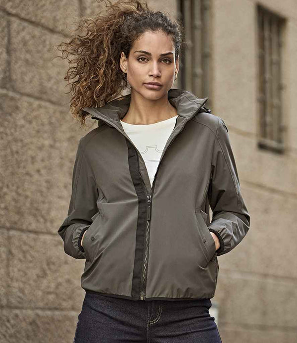 A person with curly hair wears the Tee Jays Ladies Urban Adventure Shell Jacket by Tee Jays in gray over a white shirt and dark pants, standing hands-in-pockets before a textured stone wall.
