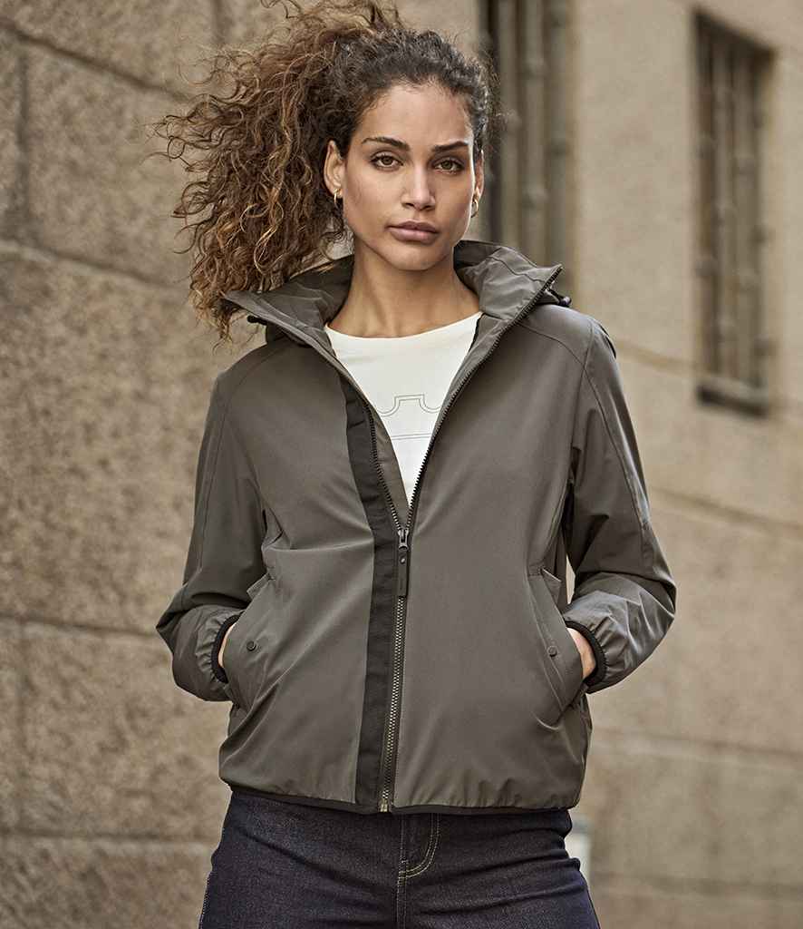 A person with curly hair wears the Tee Jays Ladies Urban Adventure Shell Jacket by Tee Jays in gray over a white shirt and dark pants, standing hands-in-pockets before a textured stone wall.