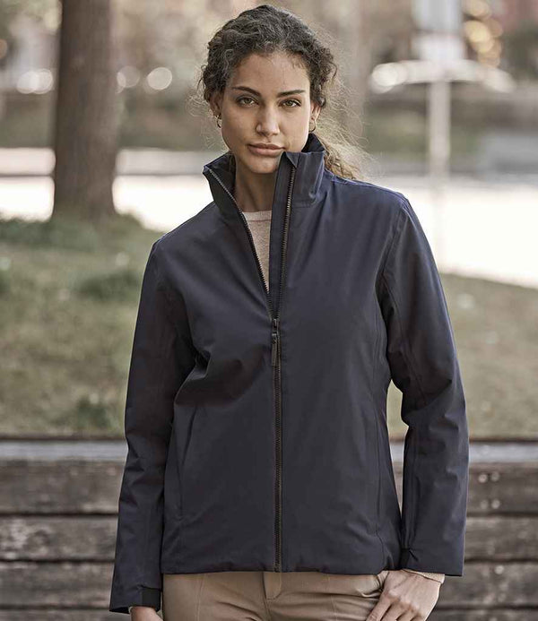 A woman with curly hair stands outdoors, wearing the Tee Jays Ladies All Weather Jacket by Tee Jays— a dark, waterproof jacket made from recycled polyester— and light-colored pants. She faces the camera neutrally, with trees and a wooden bench behind her.