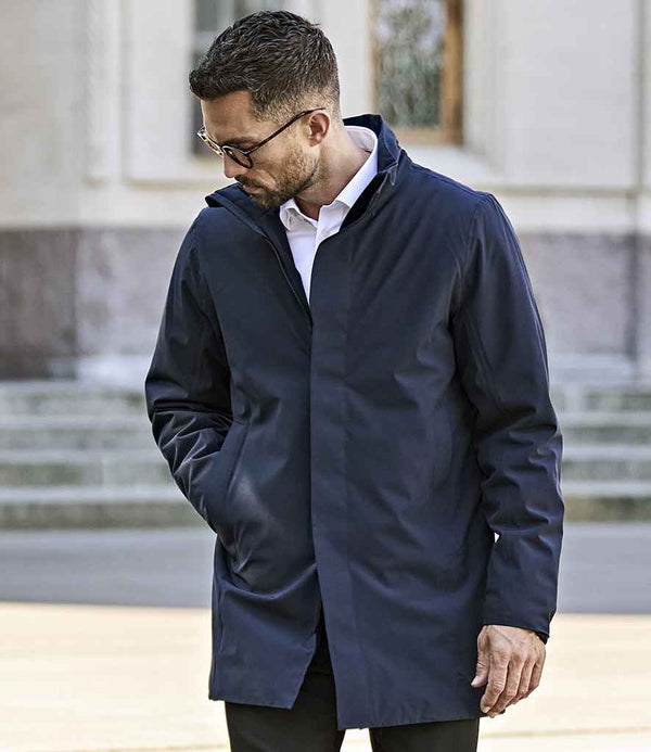 A man in glasses, a white collared shirt, and a dark blue Tee Jays All Weather Parka Jacket stands outdoors with one hand in his pocket, looking down. Stone steps and a building with windows appear in the background.