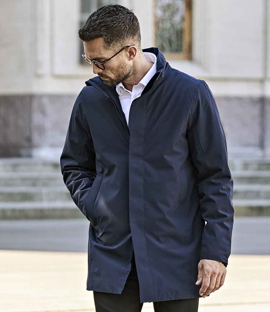 A man in glasses, a white collared shirt, and a dark blue Tee Jays All Weather Parka Jacket stands outdoors with one hand in his pocket, looking down. Stone steps and a building with windows appear in the background.