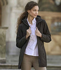 A woman stands outdoors wearing the Tee Jays Ladies All Weather Parka Jacket by Tee Jays over a white button-up shirt and beige pants, holding the jacket by its zipper. The blurred background shows some architectural details.