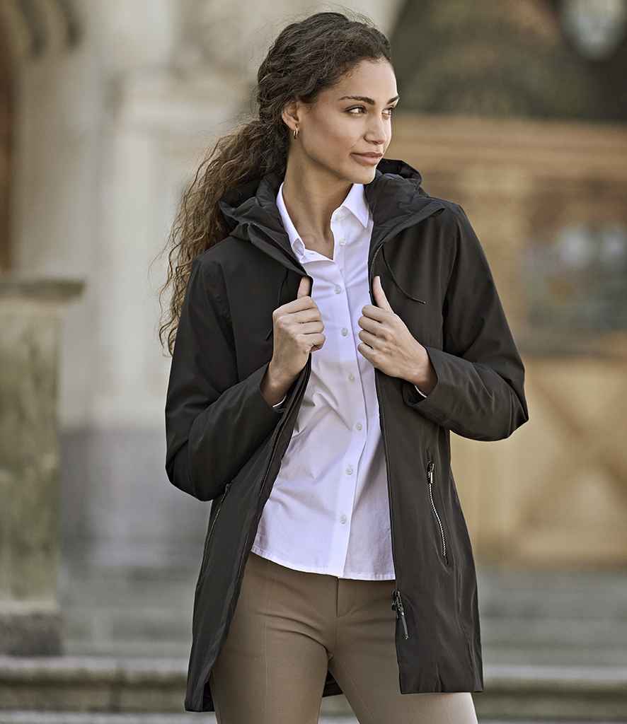 A woman stands outdoors wearing the Tee Jays Ladies All Weather Parka Jacket by Tee Jays over a white button-up shirt and beige pants, holding the jacket by its zipper. The blurred background shows some architectural details.