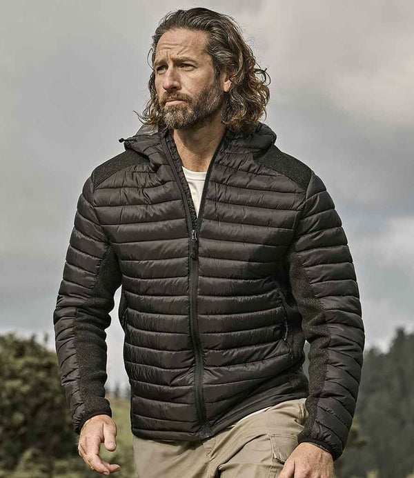 A man with long hair and a beard wears a black Tee Jays Crossover Hooded Padded Outdoor Jacket made from recycled polyester and beige pants outdoors, with trees and a cloudy sky in the background.
