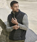 A man with short dark hair and a trimmed beard wears a Tee Jays Crossover Padded Bodywarmer—made from recycled polyester—over a light gray long-sleeve shirt. He stands outdoors, looking left, adjusting his sleeve.