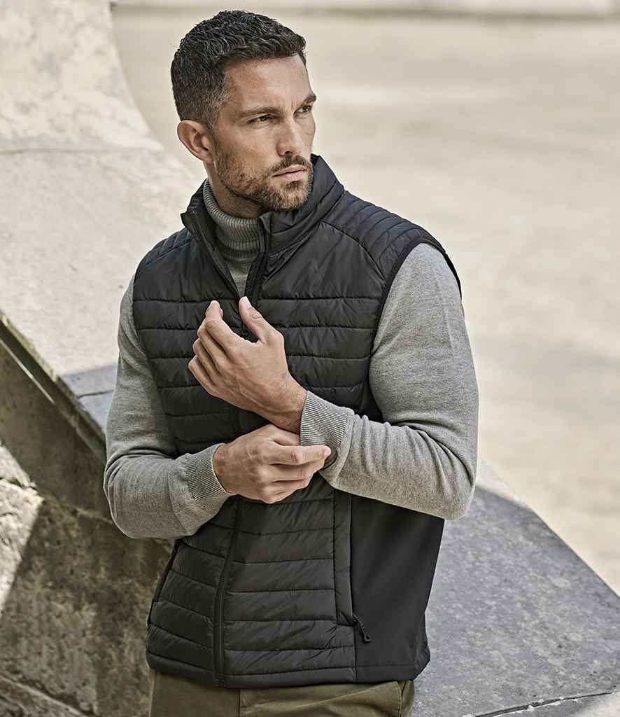 A man with short dark hair and a trimmed beard wears a Tee Jays Crossover Padded Bodywarmer—made from recycled polyester—over a light gray long-sleeve shirt. He stands outdoors, looking left, adjusting his sleeve.