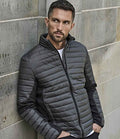 A man with short hair and a trimmed beard wears the gray Tee Jays Crossover Padded Jacket made from recycled polyester over a white shirt. He stands by a stone wall, looking sideways, one hand in his pocket.