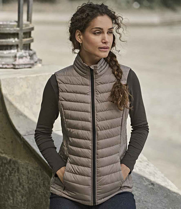 A woman with curly hair in a side braid, wearing the Tee Jays Ladies Zepelin Padded Bodywarmer over a dark long-sleeve shirt, stands outdoors with hands in pockets, gazing to the side.
