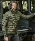 A man with curly hair and a beard wears the Tee Jays Lite Jacket by Tee Jays, an olive green showerproof jacket made from recycled nylon with warm DuPont padding, standing by an open vehicle door outdoors with trees in the background.