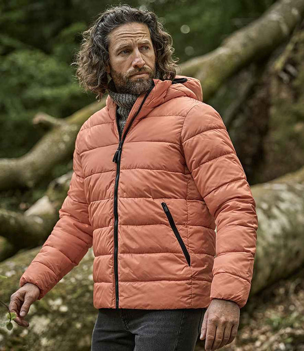 A man with wavy hair and a beard wears the Tee Jays Lite Hooded Jacket in orange by Tee Jays, featuring recycled nylon lining, along with dark pants, standing outdoors before large fallen tree trunks and green foliage.