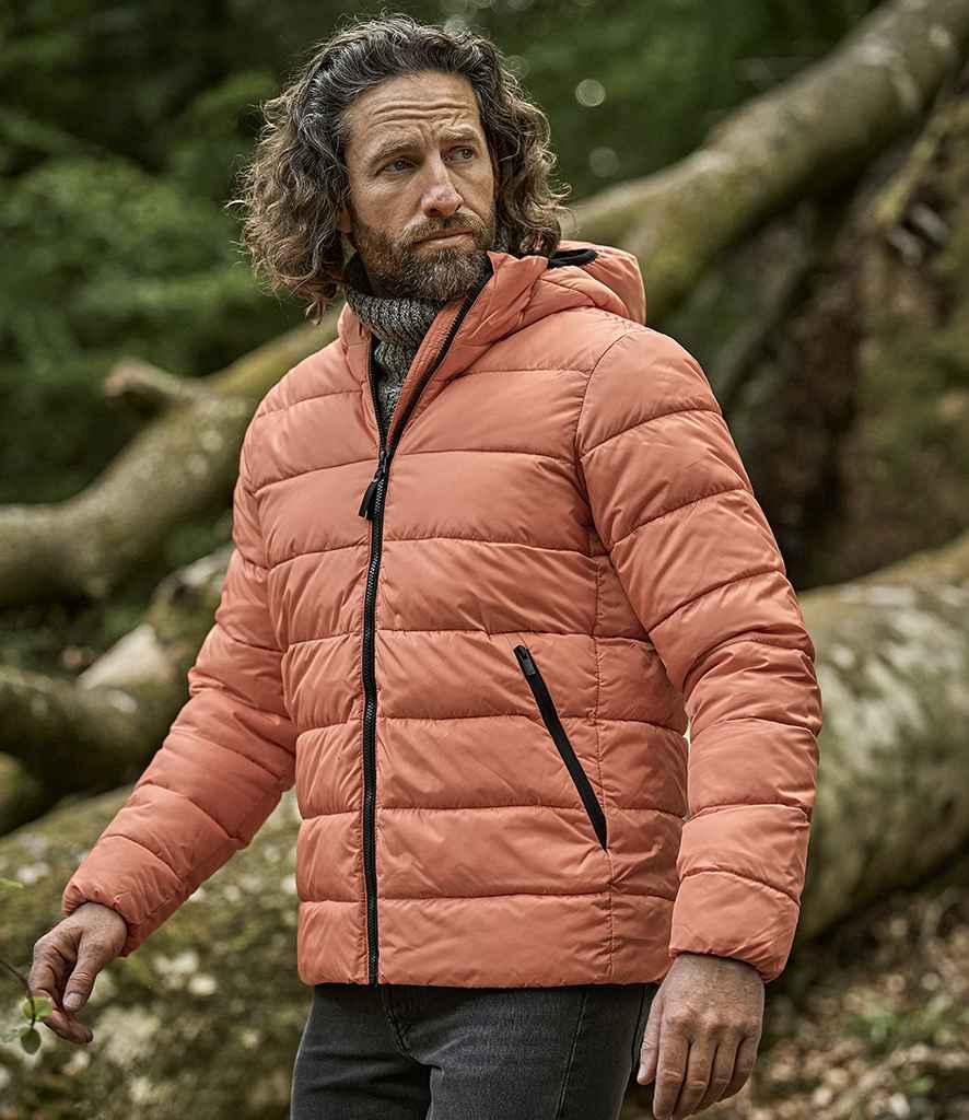 A man with wavy hair and a beard wears the Tee Jays Lite Hooded Jacket in orange by Tee Jays, featuring recycled nylon lining, along with dark pants, standing outdoors before large fallen tree trunks and green foliage.