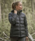 A woman wearing a Tee Jays Ladies Lite Hooded Jacket by Tee Jays stands outdoors in a forest, looking to the side with one hand adjusting her jacket collar over a turtleneck sweater.