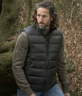 A man with long curly hair and a beard wears a gray knit sweater and the Tee Jays Lite Bodywarmer by Tee Jays. He is outdoors among moss-covered rocks and brown leaves.