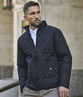 A man with short hair and a trimmed beard stands outdoors in front of a stone building, wearing the Tee Jays Richmond Jacket by Tee Jays—a dark, quilted, showerproof jacket with a classic fit and corduroy collar.