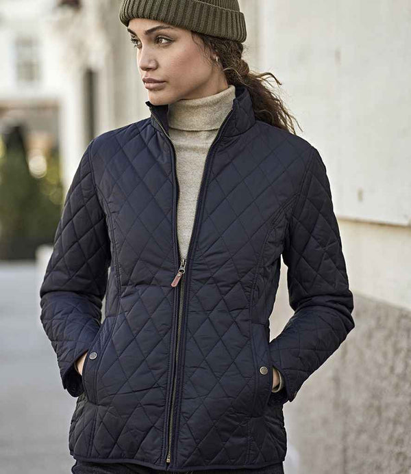 A woman stands outdoors, hands in pockets, wearing a Tee Jays Ladies Richmond Jacket with brass zip and corduroy collar, paired with a beige turtleneck and olive knit beanie. She looks to the side against a blurred backdrop.
