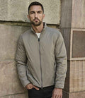 A man with short dark hair and a beard stands against a stone wall, wearing the Tee Jays PureLite Jacket by Tee Jays in light gray over a white shirt and black pants, with one hand in his pocket.