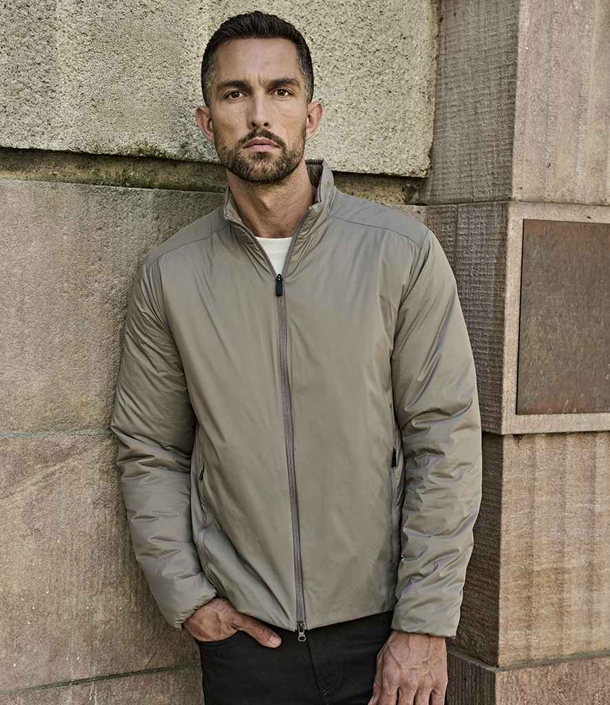 A man with short dark hair and a beard stands against a stone wall, wearing the Tee Jays PureLite Jacket by Tee Jays in light gray over a white shirt and black pants, with one hand in his pocket.