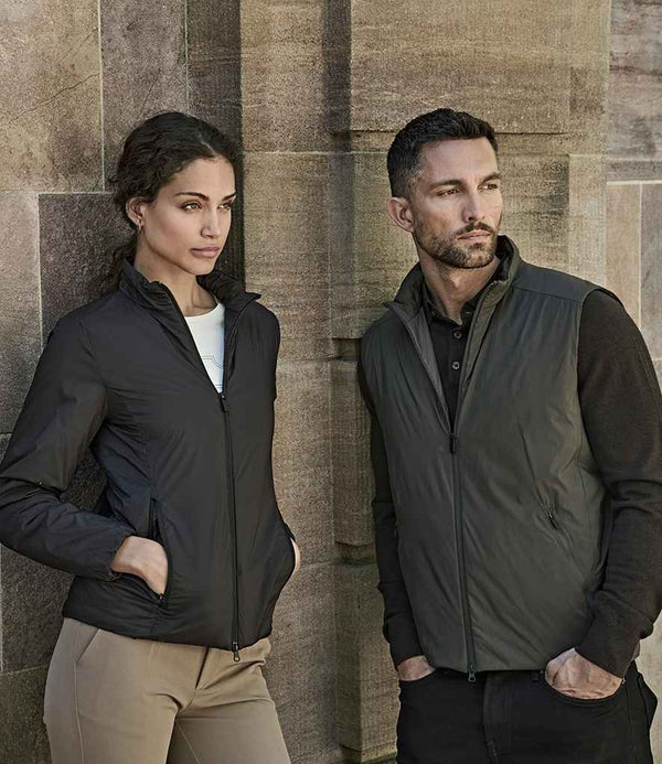 A woman in a black zip-up jacket made with DuPont™ Sorona® and beige pants stands by a stone wall. Next to her, a man wears the Tee Jays PureLite Bodywarmer by Tee Jays over a black shirt and pants. Both look away from the camera.