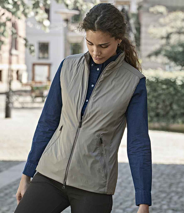 A woman with curly hair wears the Tee Jays Ladies PureLite Bodywarmer by Tee Jays in light gray over a dark blue shirt, standing outdoors on a cobblestone street with trees and buildings in the background.