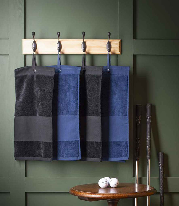 Four Towel City Deluxe Velour Golf Towels in black and navy hang from hooks on a wooden rack against a green wall; three golf clubs and two golf balls are displayed beside a round wooden table below.
